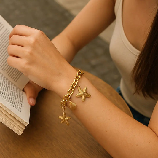 Person wearing a gold chain bracelet with starfish charms, holding an open book.