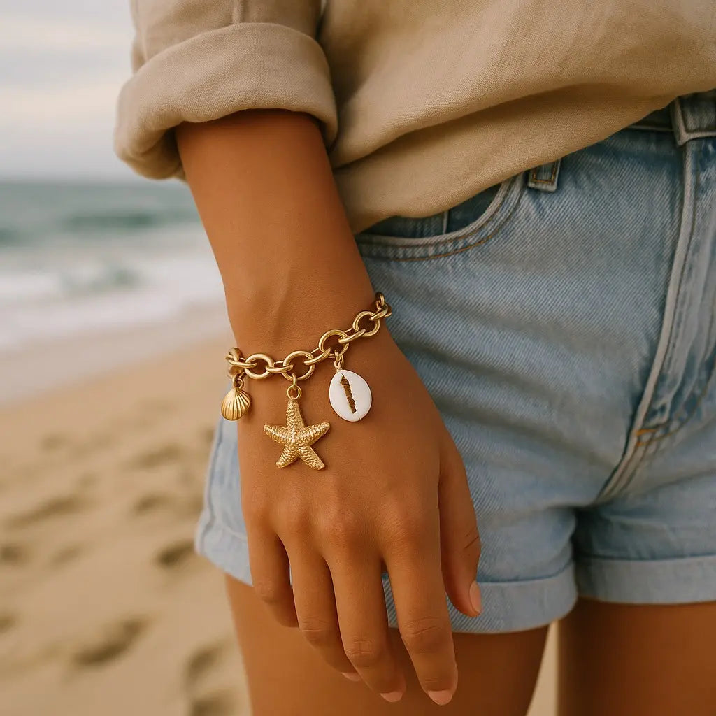 Hand wearing a gold bracelet with starfish and shell charms on a beach.