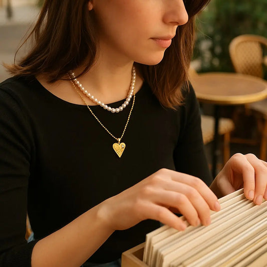Woman wearing a pearl necklace and gold heart pendant, sitting outdoors.