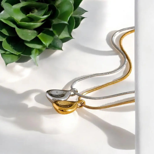 Two necklaces, one gold and one silver, on a white surface with a green plant in the background.