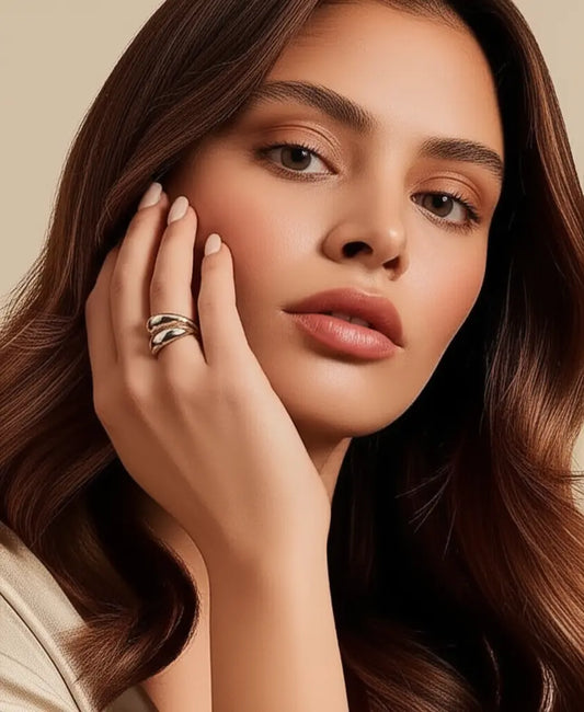 Woman with long brown hair touching her face, wearing a ring on her finger against a beige background