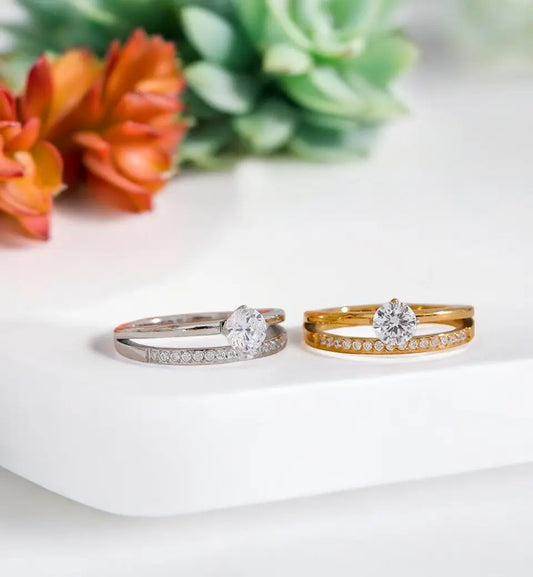 Two diamond rings, one gold and one silver, on a white surface with flowers in the background.
