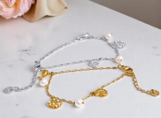 Two gold and silver anklets with charms on a marble surface.
