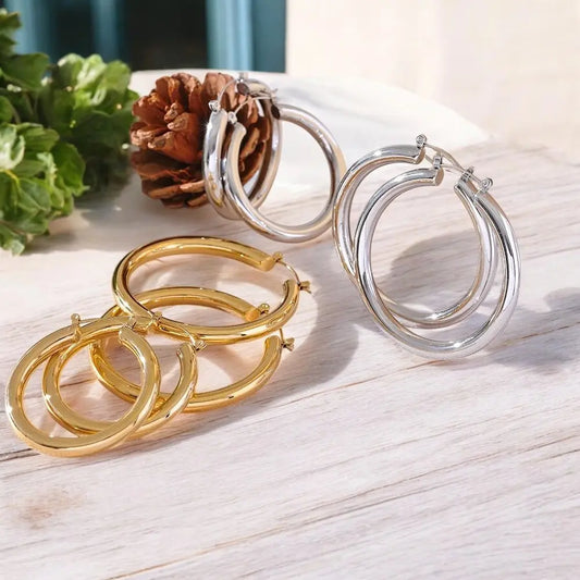 Gold and silver hoop earrings on a light wooden surface with a pine cone and green leaves.