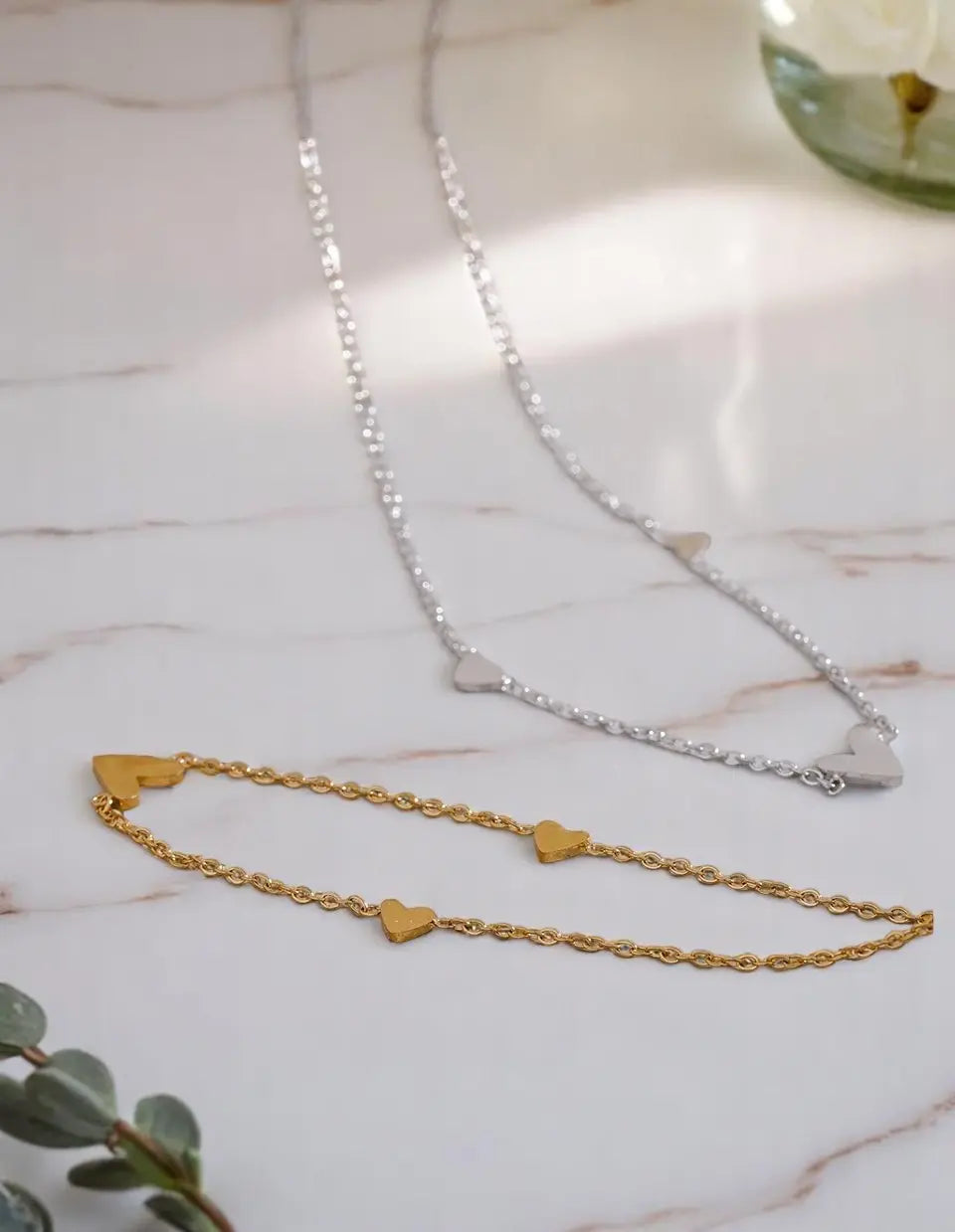 Two necklaces on a marble surface, one gold with heart pendants and one silver.