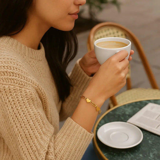 Woman holding a white mug with a drink, wearing a beige sweater and gold bracelet, sitting at a table.