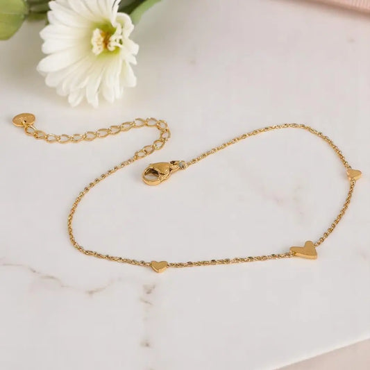 Gold anklet with heart-shaped charms on a marble surface with a white flower.