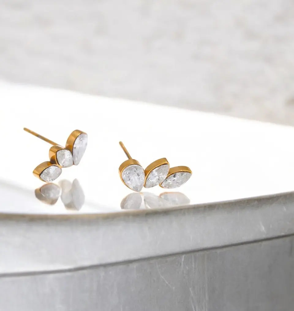 Gold and clear crystal stud earrings on a reflective surface with a light background
