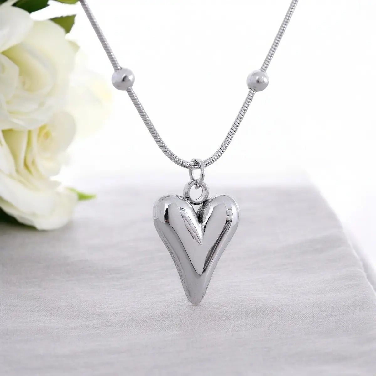 Silver heart-shaped pendant necklace on a white background with white flowers.
