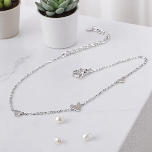 Silver heart-shaped anklet with matching earrings on a marble surface with potted plants.