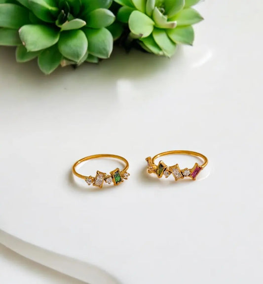 Two gold rings with gemstones on a white surface with green succulents in the background
